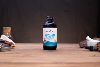 A bottle of Nordic Naturals Omega-3 dietary supplement placed on a wooden tabletop with blurred natural and kitchen-style props in the background.