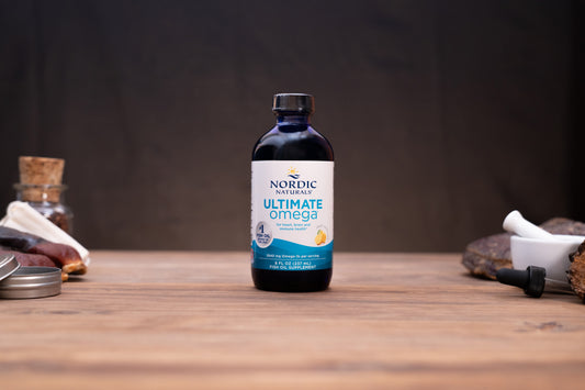 A bottle of Nordic Naturals Omega-3 dietary supplement placed on a wooden tabletop with blurred natural and kitchen-style props in the background.