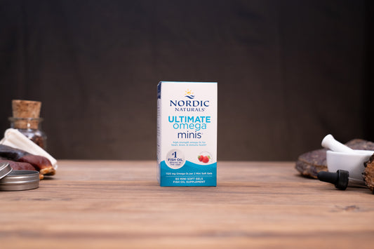 Nordic Naturals Ultimate Omega Minis supplement box standing on a wooden tabletop with blurred jars and a white mortar in the background.