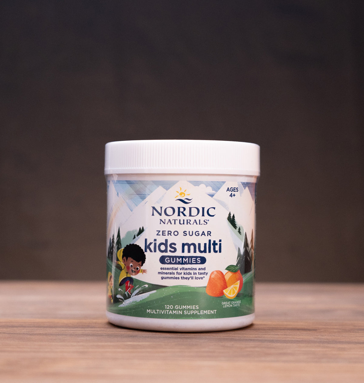 A jar of Nordic Naturals Kids Multi supplement powder stands on a wooden table against a dark background, with small natural props like dried herbs, a cork stopper, and a white mortar and pestle blurred in the background.