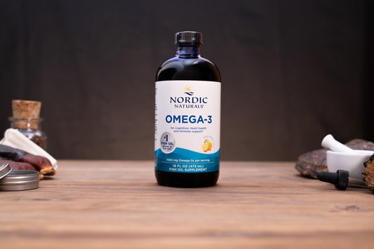 A bottle of Nordic Naturals Omega-3 dietary supplement standing on a wooden tabletop against a dark background, with small props visible on both sides.