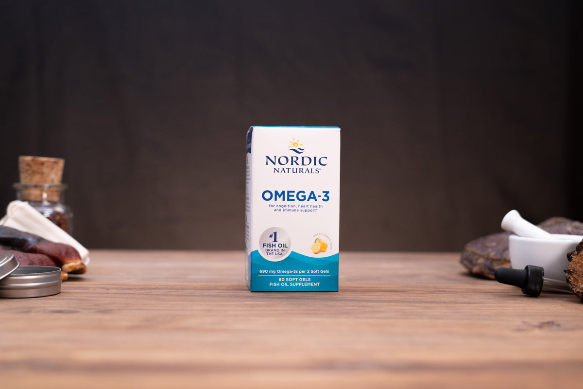 A box of Nordic Naturals Omega-3 fish oil supplement stands centered on a wooden tabletop against a dark background, with small blurred props on both sides.