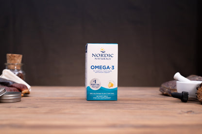 A box of Nordic Naturals Omega-3 fish oil supplement stands centered on a wooden tabletop against a dark background, with small blurred props on both sides.