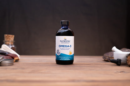 A bottle of Nordic Naturals Omega-3 dietary supplement placed on a wooden tabletop with blurred natural and kitchen-style props in the background.