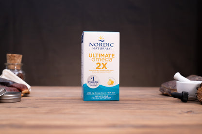 Nordic Naturals Ultimate Omega 2X fish oil supplement box standing on a wooden tabletop with blurred natural props in the background.