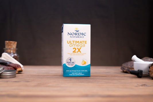 Nordic Naturals Ultimate Omega 2X fish oil supplement box standing on a wooden tabletop with blurred natural props in the background.