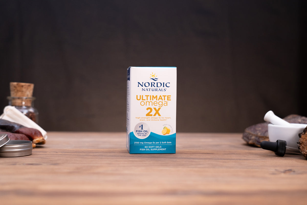 Nordic Naturals Ultimate Omega 2X dietary supplement box standing on a wooden tabletop with blurred jars and a white mortar in the background.