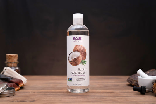A bottle of Now Foods liquid coconut oil stands on a wooden surface against a dark background, with small kitchen or craft props visible on both sides.