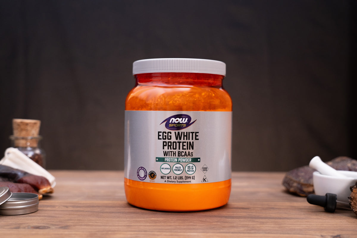 A jar of Now Foods Egg White Protein with BCAAs sits on a wooden table against a dark background, surrounded by small rustic props like a cork stopper, a small bottle, and a mortar and pestle.