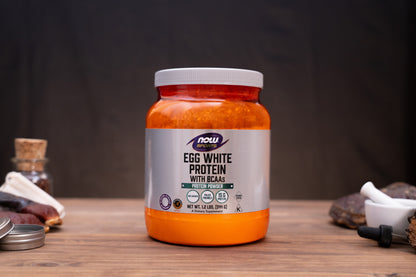 A jar of Now Foods Egg White Protein with BCAAs sits on a wooden table against a dark background, surrounded by small rustic props like a cork stopper, a small bottle, and a mortar and pestle.