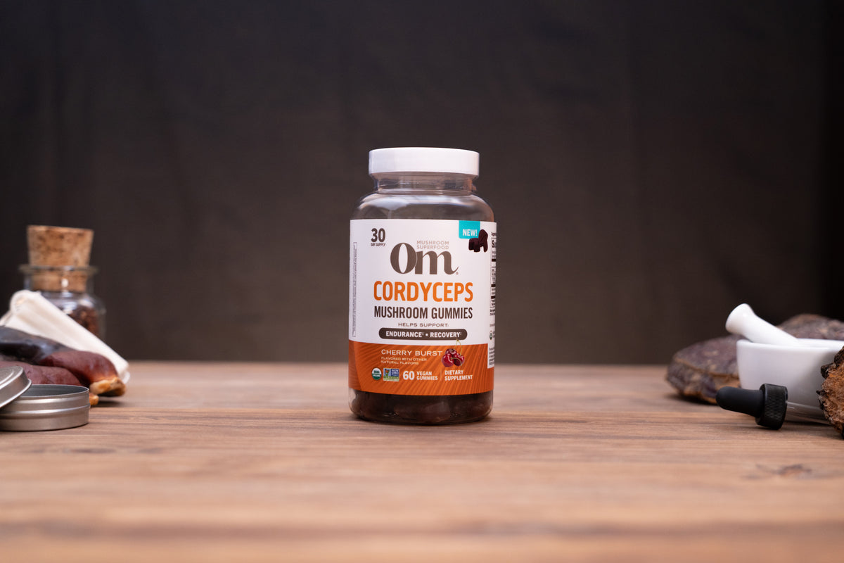 A bottle of Om Cordyceps Mushroom Gummies stands on a wooden tabletop against a dark backdrop, with small rustic props visible on both sides.