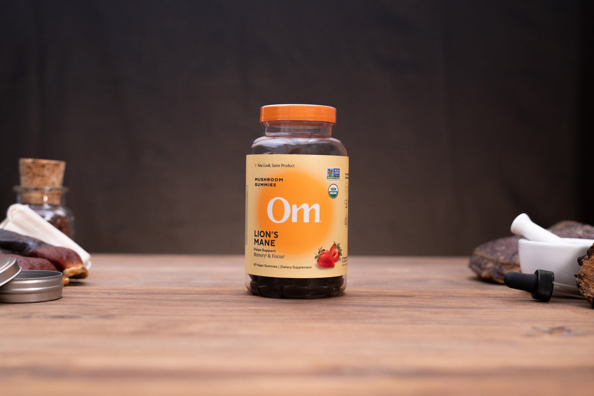 A bottle of Om Lion's Mane supplement stands on a wooden table against a dark background, with small herbal and kitchen props blurred on both sides.