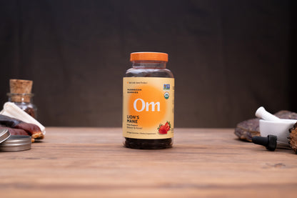 A bottle of Om Lion's Mane supplement stands on a wooden table against a dark background, with small herbal and kitchen props blurred on both sides.