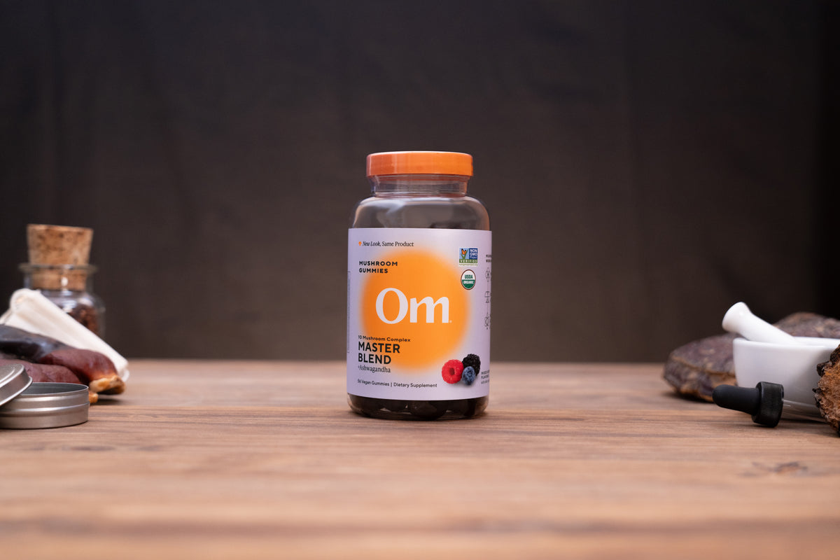 A bottle of Om Master Blend herbal supplement centered on a wooden table, with blurred natural props on both sides against a dark background.