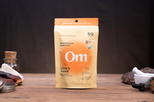 A stand-up pouch of Om Mushroom Powder in Lion's Mane flavor, standing on a wooden surface with rustic herbal props in the background.