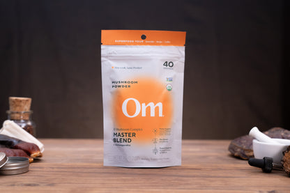 A stand-up pouch of Om Mushroom Powder Master Blend standing on a wooden surface, with a dark blurred background and small kitchen/spice props around it.