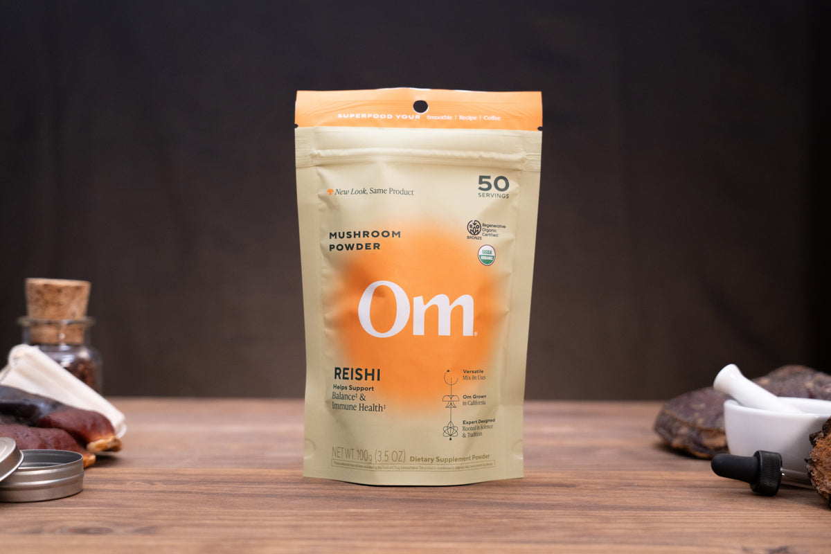 A stand-up pouch of Om Reishi Mushroom Powder centered on a wooden surface, with a dark blurred background and small surrounding props including jars, stones, and a dropper.
