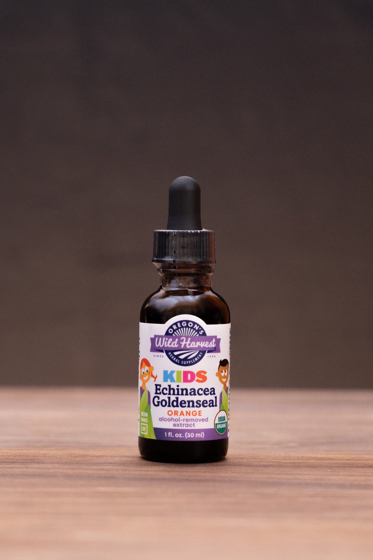 A small amber glass dropper bottle labeled 'Kids Immunity Goldenseal' stands centered on a wooden tabletop against a dark backdrop, with blurred herbal props on both sides.