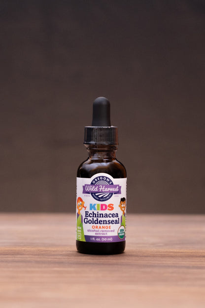 A small amber glass dropper bottle labeled 'Kids Immunity Goldenseal' stands centered on a wooden tabletop against a dark backdrop, with blurred herbal props on both sides.