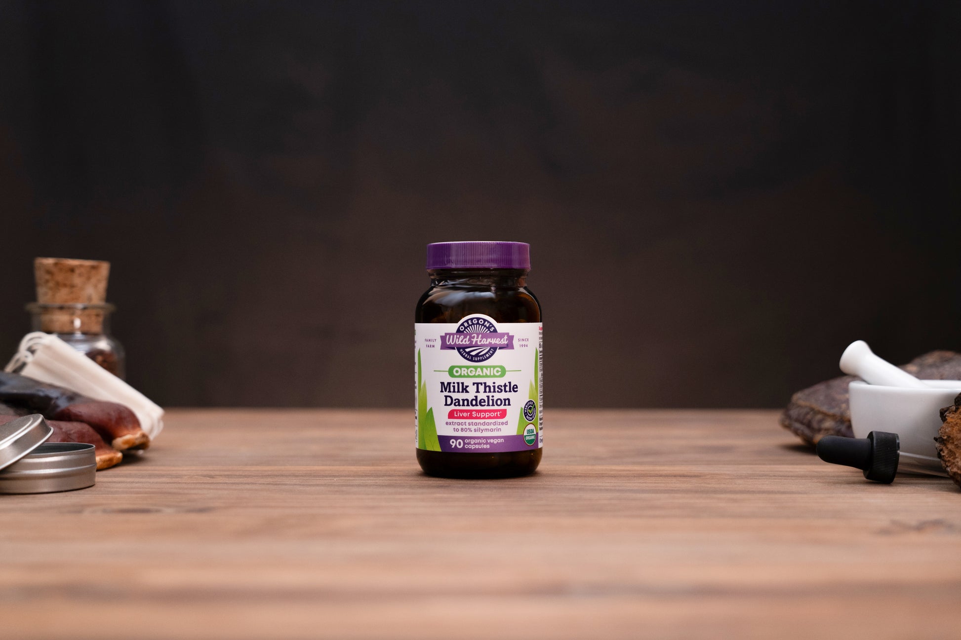 A bottle of Organic Apothecary Milk Thistle Dandelion supplement centered on a wooden tabletop, with blurred herbal props on either side against a dark background.