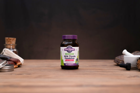 A bottle of Organic Apothecary Milk Thistle Dandelion supplement centered on a wooden tabletop, with blurred herbal props on either side against a dark background.