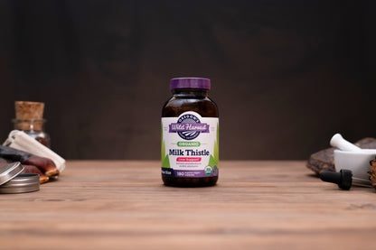 A bottle of Mother Nature's Milk Thistle dietary supplement stands on a wooden tabletop against a dark, blurred background. The bottle is amber brown with a purple cap and a white, purple, and green label.