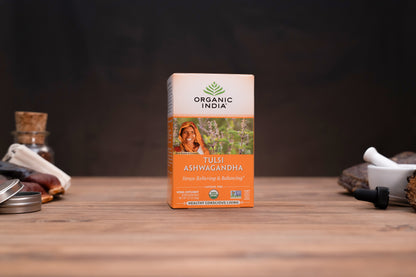 A box of Organic India Tulsi Ashwagandha tea stands centered on a wooden tabletop, with blurred herbal props on both sides against a dark background.