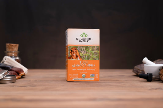 A box of Organic India Tulsi Ashwagandha tea stands centered on a wooden tabletop, with blurred herbal props on both sides against a dark background.