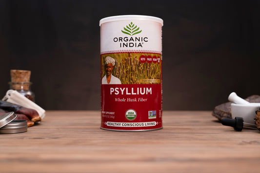 A canister of Organic India Psyllium Whole Husk Fiber standing on a wooden tabletop, with small kitchen or herbal-prep props visible on both sides against a dark background.