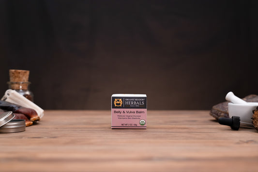A small boxed product labeled 'The Herbalist Baby & Vena Barn' sits centered on a wooden tabletop against a dark brown backdrop, with blurred herbal props on both sides.