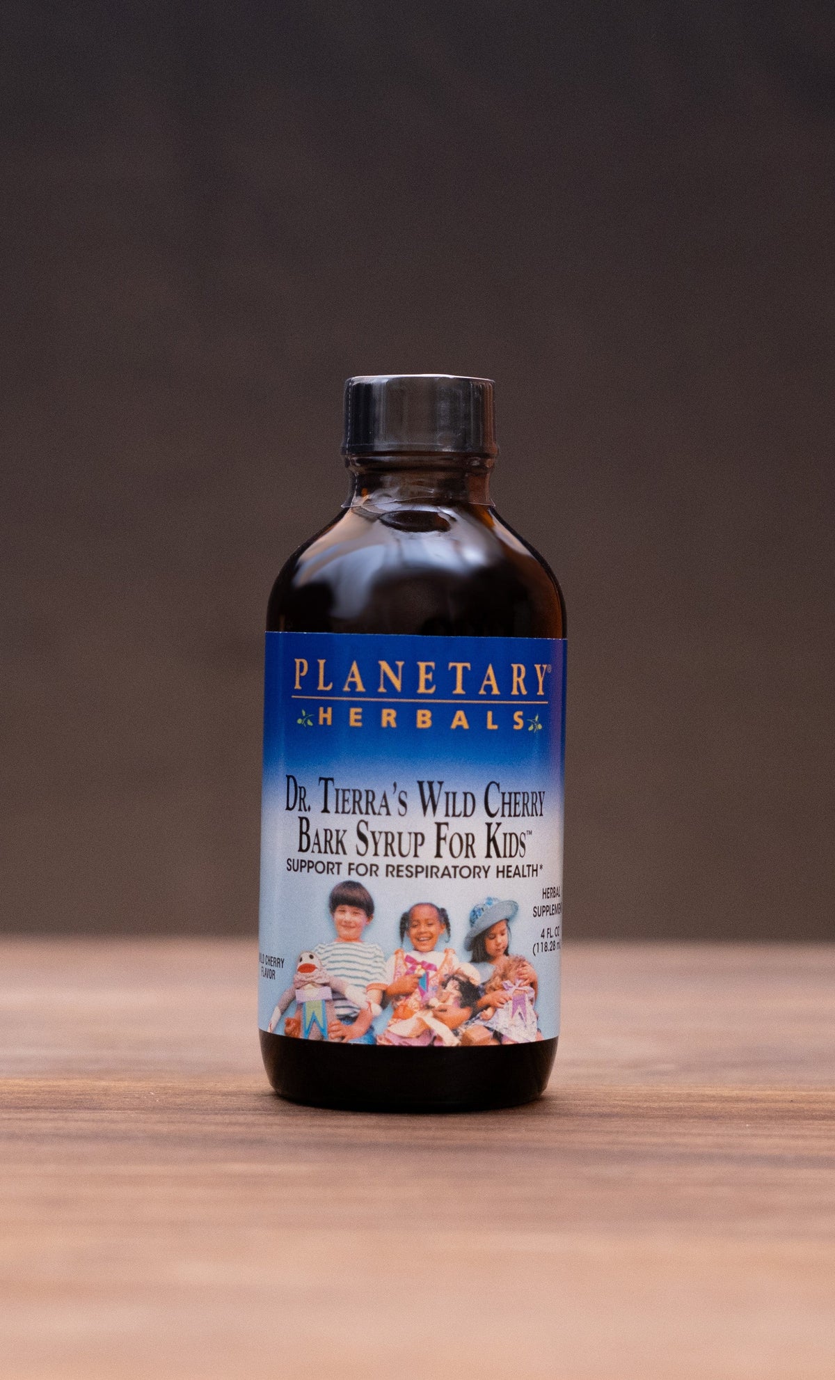 A small amber glass bottle labeled 'Planetary Herbs' sits centered on a wooden surface against a dark backdrop, with blurred herbal props on both sides.