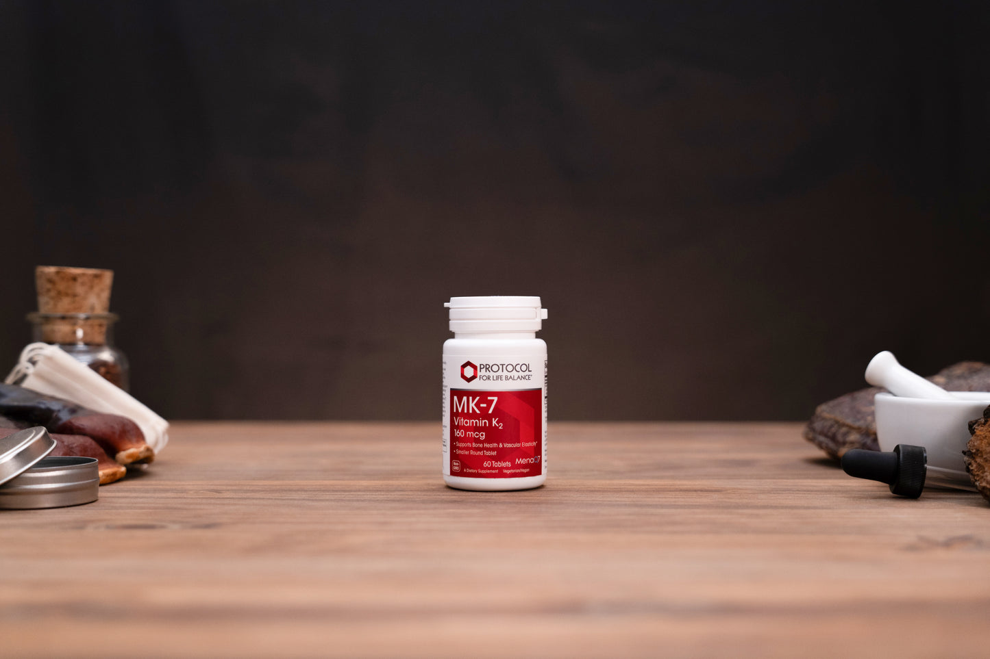 A white bottle of Morinocoo MK-7 Vitamin K2 1000 IU stands centered on a wooden tabletop, flanked by blurred herbal and apothecary props against a dark brown background.