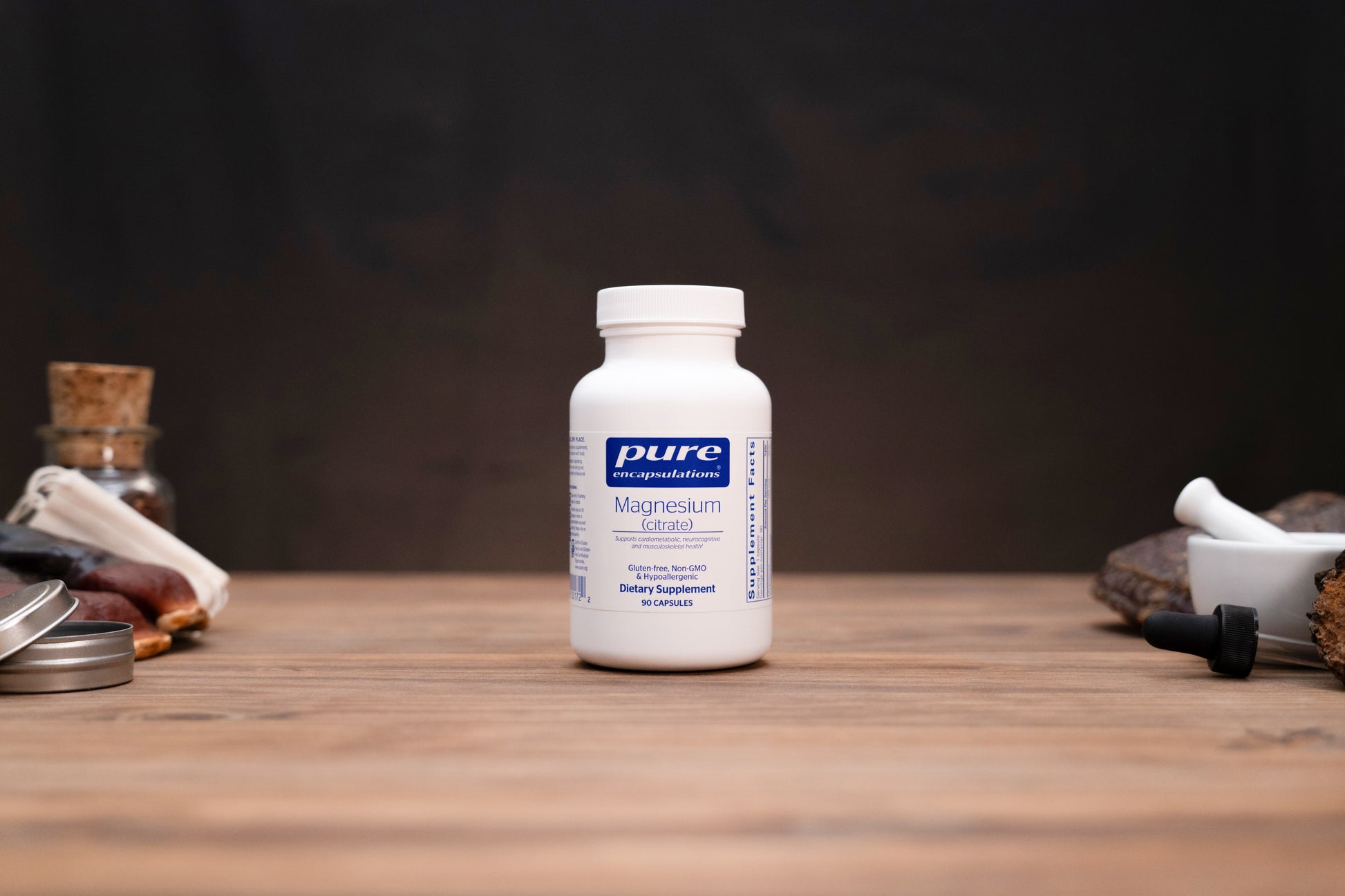 A white bottle of Pure Encapsulations Magnesium Glycine supplement stands on a wooden tabletop against a dark, moody background, styled with herbal props on both sides.