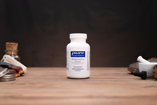 A white bottle of Pure Encapsulations Magnesium Glycine supplement stands on a wooden tabletop against a dark, moody background, styled with herbal props on both sides.