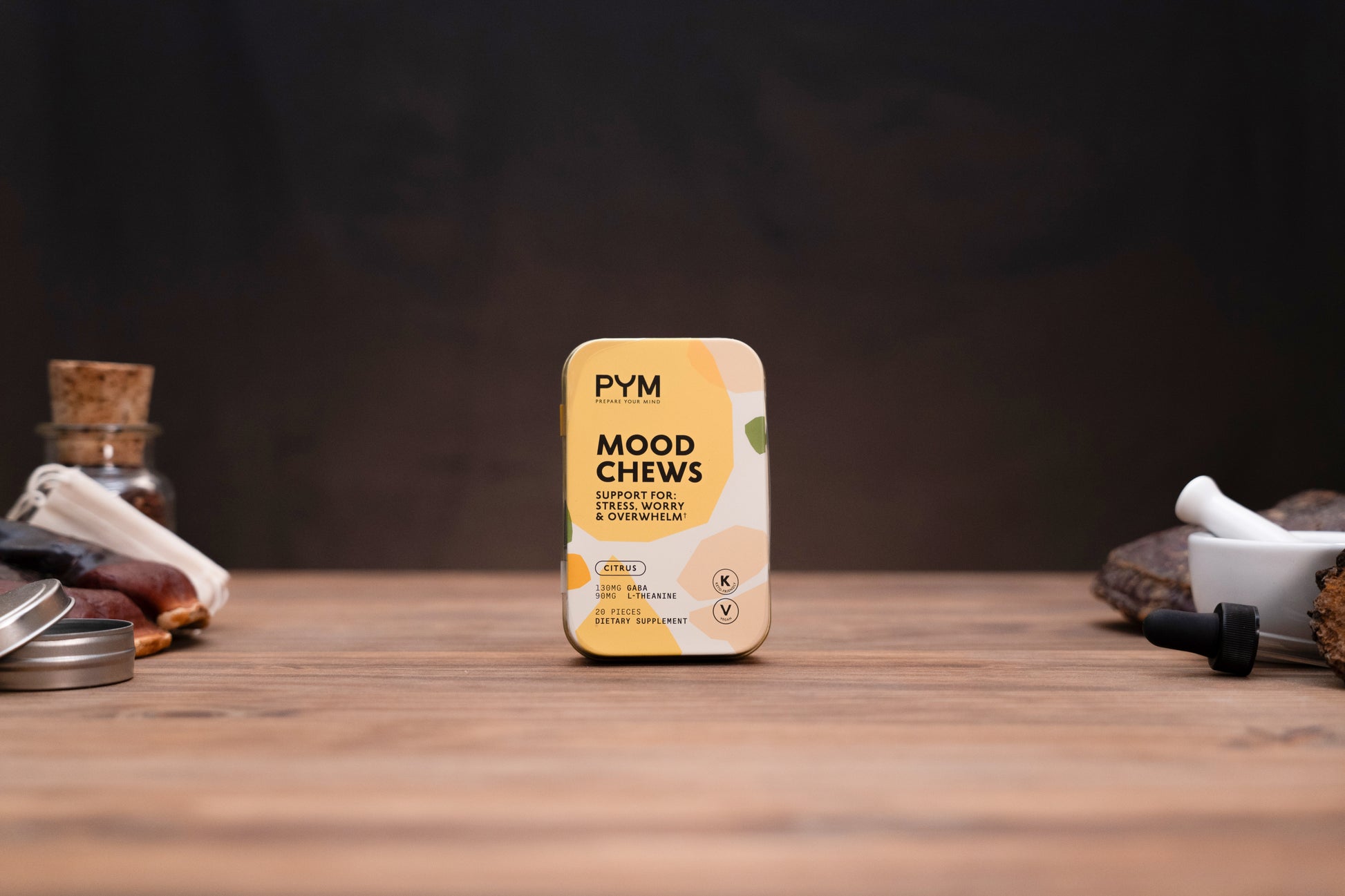 A tin of PYM Mood Chews, flavoring notes: Cinnamon, Cloves & Vanilla, centered on a wooden tabletop with a dark, moody background and styled herbal props on either side.