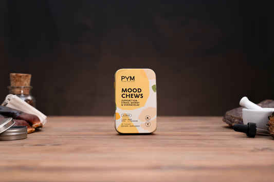 A tin of PYM Mood Chews, flavoring notes: Cinnamon, Cloves & Vanilla, centered on a wooden tabletop with a dark, moody background and styled herbal props on either side.