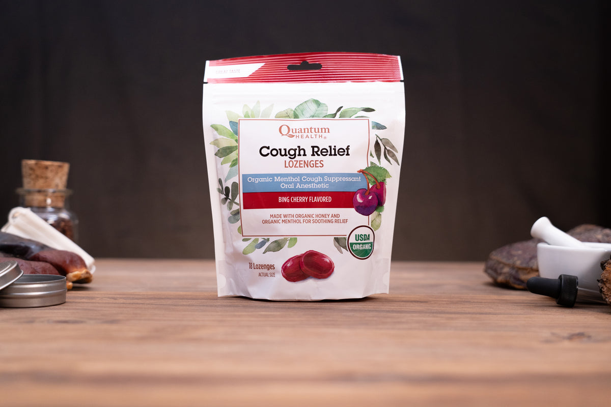 A stand-up pouch of Quantum Health Cough Relief Lozenge in Bing Cherry Flavored, displayed on a wooden surface with blurred herbal and mortar-and-pestle props in the background.