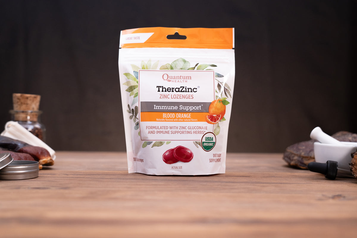 A stand-up pouch of Quantum Health TheraZinc Zinc Glucone in Blood Orange flavor, displayed on a wooden tabletop with rustic herbal props in the background.