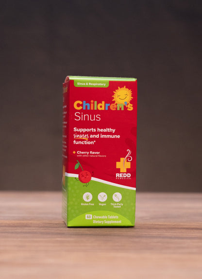 A box of Child Sinus syrup for children stands centered on a wooden tabletop against a dark, blurred background. The packaging is red and green with yellow text and graphics.