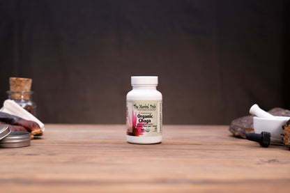 A white bottle of The Herbal Park Organic Chaga supplement stands centered on a wooden tabletop, with blurred herbal props on both sides against a dark backdrop.