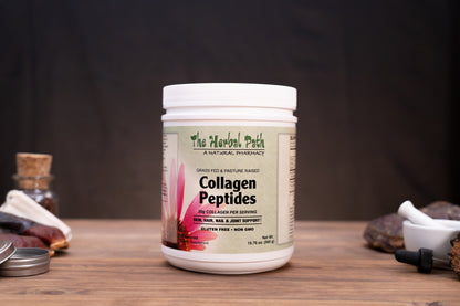 A white plastic jar of The Herbal Path Collagen Peptides supplement, displayed on a wooden table with natural props like a cork stopper, dried roots, and a mortar and pestle against a dark background.