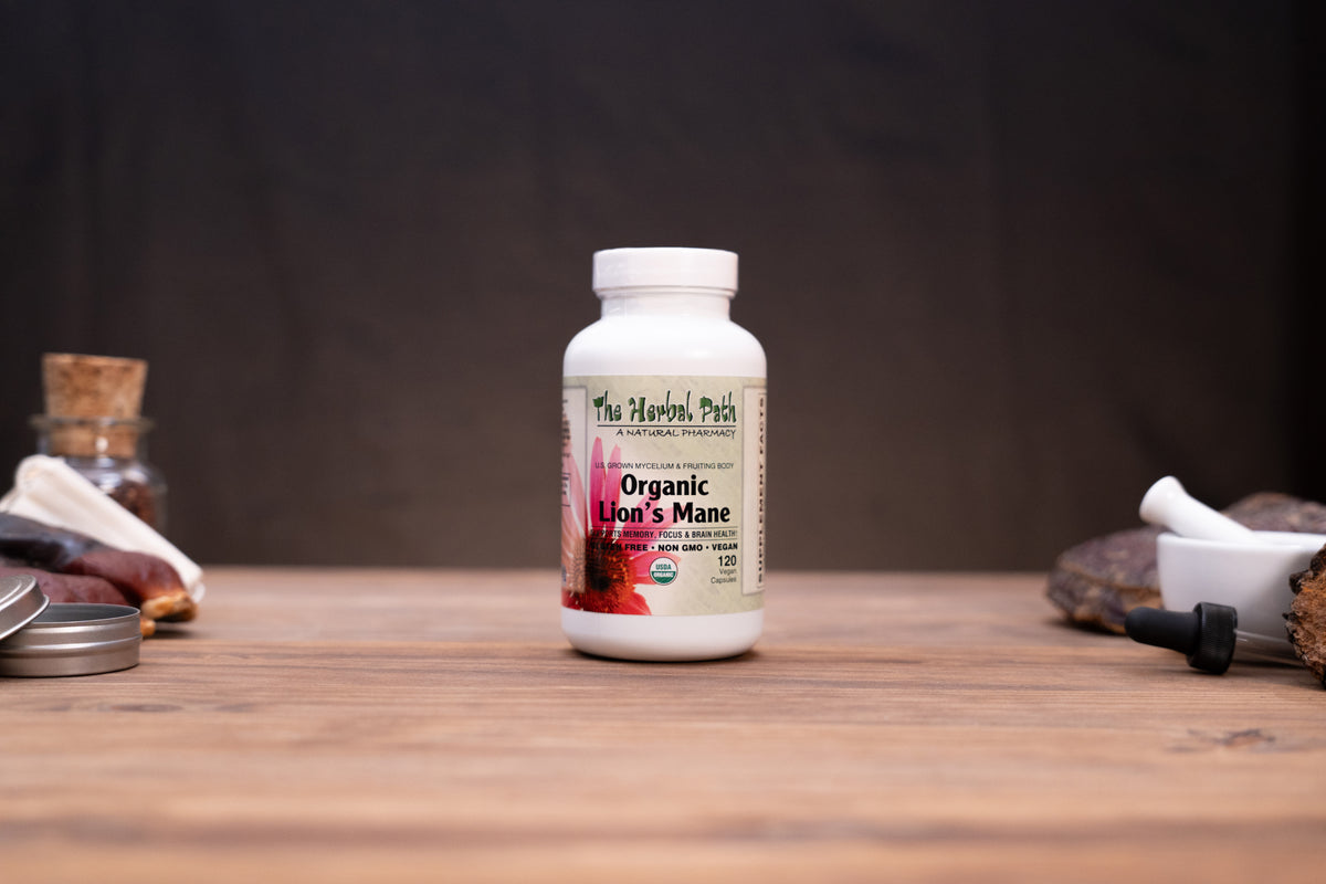 A white bottle of The Herbal Patch Organic Lion's Mane dietary supplement, standing on a wooden tabletop with rustic herbal props blurred in the background.