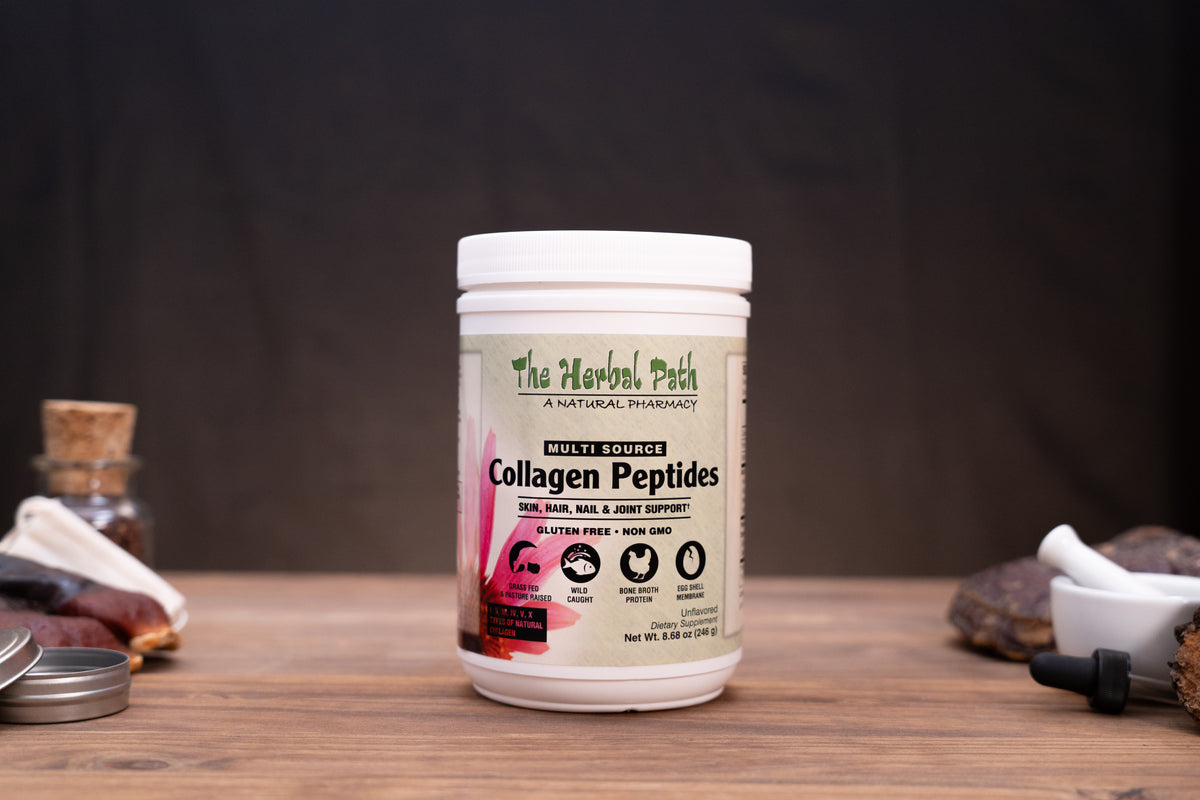 A white plastic tub of The Herbal Path Collagen Peptides centered on a wooden table, with a dark draped backdrop behind it and small rustic props on both sides.