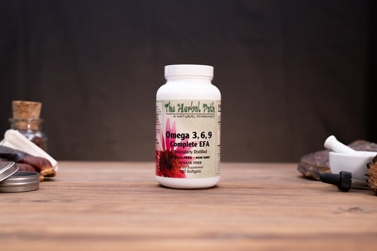 A white plastic bottle of 'The Harvest Path' Omega 3, 6, 9 Complete EFA dietary supplement, standing on a wooden surface with blurred natural elements in the background.