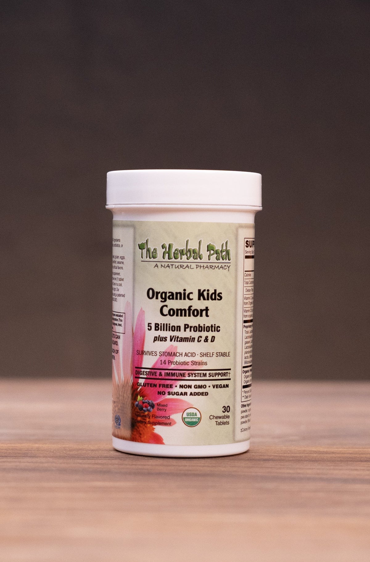 A white plastic bottle of The Herbal Patch Organic Kids Lemonade powder sits on a wooden tabletop against a dark background, surrounded by blurred herbal-themed props.