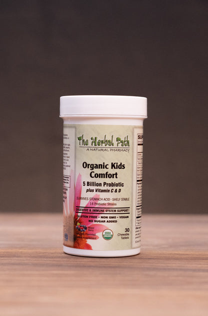 A white plastic bottle of The Herbal Patch Organic Kids Lemonade powder sits on a wooden tabletop against a dark background, surrounded by blurred herbal-themed props.