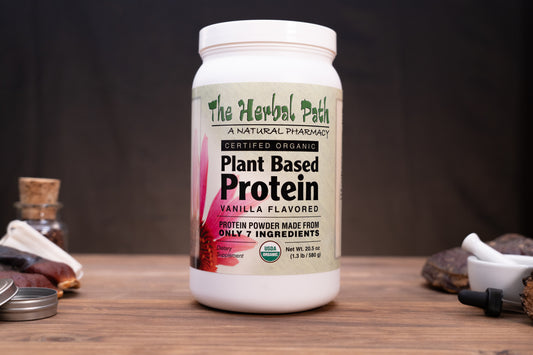 A white plastic tub of plant-based protein powder by The Herbal Path, labeled Vanilla Flavored, displayed on a wooden surface with herbal props in the background.
