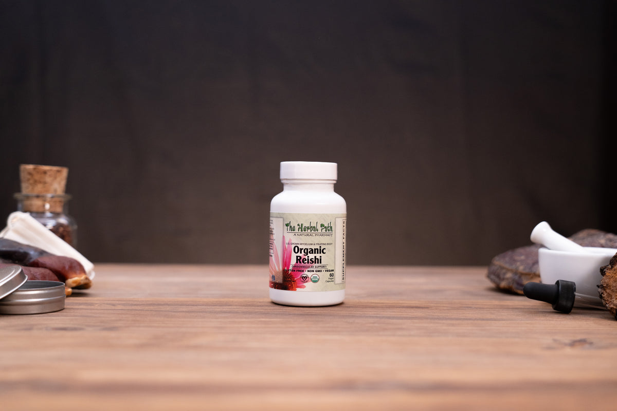 A white bottle of The Herbal Park Organic Chaga supplement stands centered on a wooden tabletop, with blurred herbal props on both sides against a dark backdrop.