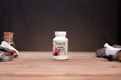 A white bottle of The Herbal Path Organic Turkey Tail supplement standing on a wooden tabletop against a dark backdrop, with rustic props visible on both sides.