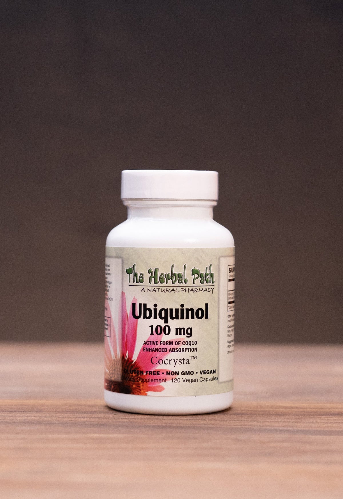 A white bottle of The Herbal Park Organic Chaga supplement stands centered on a wooden tabletop, with blurred herbal props on both sides against a dark backdrop.